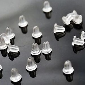 May include: A selection of clear, soft plastic earring backs with black posts. The earring backs are scattered across a reflective, dark surface, highlighting their translucent nature and simple design. These are essential jewellery components.