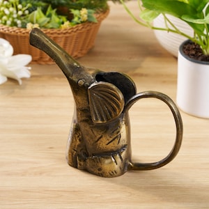 Peut inclure: Un arrosoir en forme d'éléphant de couleur bronze. L'éléphant a une longue trompe, de grandes oreilles et une poignée circulaire. L'arrosoir est en métal et a un aspect vintage. Le fond comprend des plantes et une surface en bois.