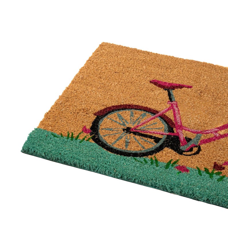 K&ouml;nnte beinhalten: Rechteckige Fu&szlig;matte mit einem Fahrrad-Design. Das Fahrrad ist rosa mit einem roten Rad und vor einem beigen Hintergrund mit gr&uuml;nem Gras und rosa Blumen. Der Rand der Matte ist gr&uuml;n.