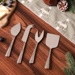May include: A collection of four silver cheese knives and serving utensils. The set includes a flat cheese knife, a curved cheese knife, a cheese fork, and a spatula. The handles have a textured design. The utensils are arranged on a wooden surface.