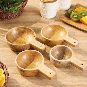 May include: A set of four wooden measuring cups with handles. The cups are labeled 1 Cup, 1/2 Cup, 1/3 Cup, and 1/4 Cup. The cups are made of light-colored wood and are arranged on a wooden surface.