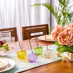 May include: Four colorful glass incense holders in yellow, purple, green, and pink, each with a red incense stick. The holders are square-shaped and sit on a light blue table runner. A bouquet of pink flowers and a bowl of grapes are in the background.