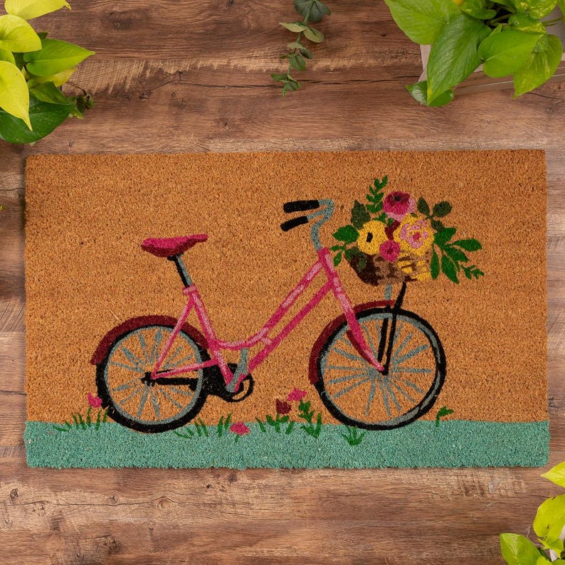 K&ouml;nnte beinhalten: Rechteckige Fu&szlig;matte mit einem Design eines rosa Fahrrades mit einem Blumenkorb. Die Matte hat einen braunen Hintergrund und einen gr&uuml;nen Rand. Das Fahrrad ist etwa 76 cm lang.