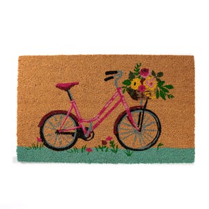 K&ouml;nnte beinhalten: Eine rechteckige Fu&szlig;matte mit einem Design eines rosa Fahrrades, das einen Blumenkorb tr&auml;gt. Das Fahrrad hat blaue R&auml;der und einen roten Sattel. Der Hintergrund ist beige mit einem gr&uuml;nen Rand unten. Die Matte besteht aus Naturfaser.
