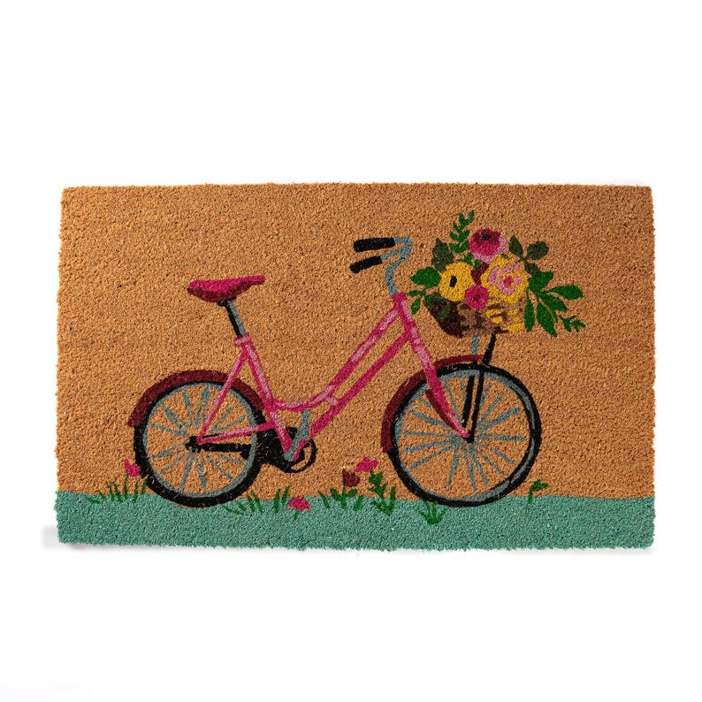 K&ouml;nnte beinhalten: Eine rechteckige Fu&szlig;matte mit einem Design eines rosa Fahrrades, das einen Blumenkorb tr&auml;gt. Das Fahrrad hat blaue R&auml;der und einen roten Sattel. Der Hintergrund ist beige mit einem gr&uuml;nen Rand unten. Die Matte besteht aus Naturfaser.