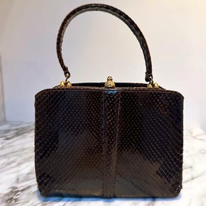 Può includere: Una borsa vintage marrone con motivo a pelle di serpente. La borsa rettangolare ha un manico curvo e finiture color oro. La borsa si chiude con una chiusura e ha una finitura lucida. La borsa è appoggiata su una superficie di marmo.