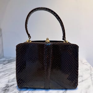 Puede incluir: Un bolso de mano vintage marrón con un estampado de piel de serpiente. El bolso estructurado tiene un asa curva y herrajes dorados. El cierre está centrado en la parte superior y el bolso parece estar en buen estado.