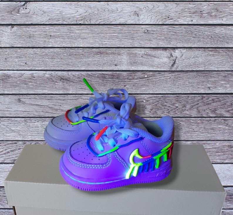 Puede incluir: Un par de zapatillas de deporte de color morado para ni&ntilde;os peque&ntilde;os con detalles coloridos y un logotipo swoosh de colores arco&iacute;ris. Las zapatillas tienen una entresuela blanca y una suela exterior azul claro.