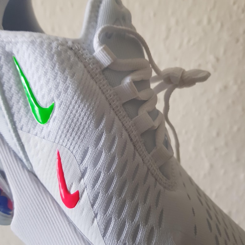 May include: Close-up of a white athletic shoe with a textured pattern. The shoe features a green Nike swoosh on the side and a red swoosh near the toe. White laces are visible.