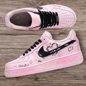 May include: Pink Air Force 1 sneakers with black laces and a black Nike swoosh. The shoes feature hand-drawn heart and doodle designs. The sole has a pink gradient design.
