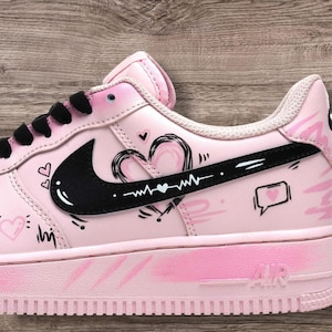 May include: Pink Air Force 1 sneakers with black accents and heart-themed illustrations. The sneakers feature a black Nike swoosh, black laces, and a pink sole. The design includes hearts, speech bubbles, and a heartbeat line.