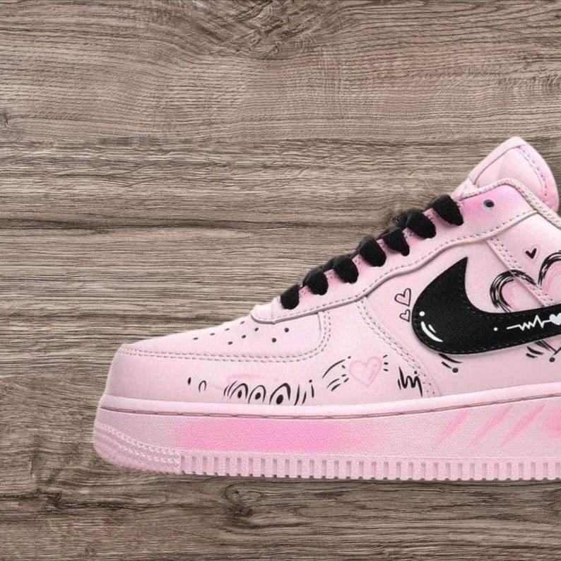 May include: Pink and white Air Force 1 sneakers with black laces and a black Nike swoosh. The shoe features hand-drawn heart and line art details. The sole is pink.