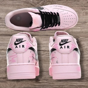 May include: Pink Nike Air Force 1 sneakers with black laces and accents. The shoes feature the Nike Air logo on the heel and decorative pink floral designs. The shoes are displayed on a wooden surface.