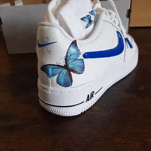 Może przedstawiać: Białe sk&oacute;rzane buty sportowe z niebieskim znaczkiem i niebieskim motylem namalowanym na boku. Słowo "AIR" jest nadrukowane na podeszwie.