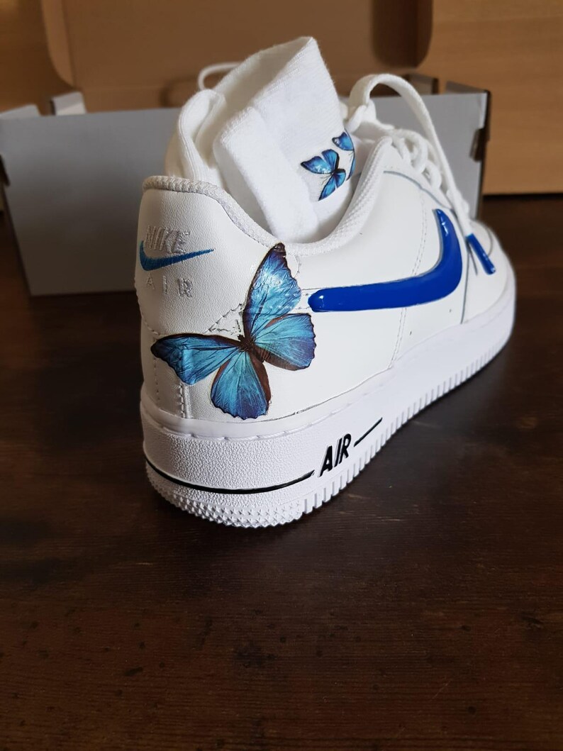 Może przedstawiać: Białe sk&oacute;rzane buty sportowe z niebieskim znaczkiem i niebieskim motylem namalowanym na boku. Słowo "AIR" jest nadrukowane na podeszwie.