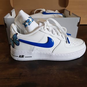 Może przedstawiać: Białe sk&oacute;rzane buty sportowe o niskim profilu z niebieskim znaczkiem i niebieskimi wzorami motyli. Słowo "AIR" jest nadrukowane na boku buta.