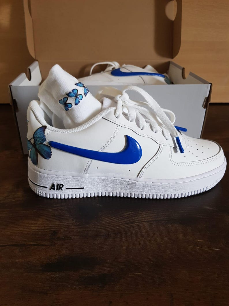 Może przedstawiać: Białe sk&oacute;rzane buty sportowe o niskim profilu z niebieskim znaczkiem i niebieskimi wzorami motyli. Słowo "AIR" jest nadrukowane na boku buta.