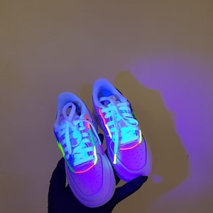 Puede incluir: Un par de zapatillas blancas con detalles en verde ne&oacute;n, rosa y azul. Las zapatillas est&aacute;n iluminadas con luz negra, lo que hace que los colores ne&oacute;n brillen intensamente.