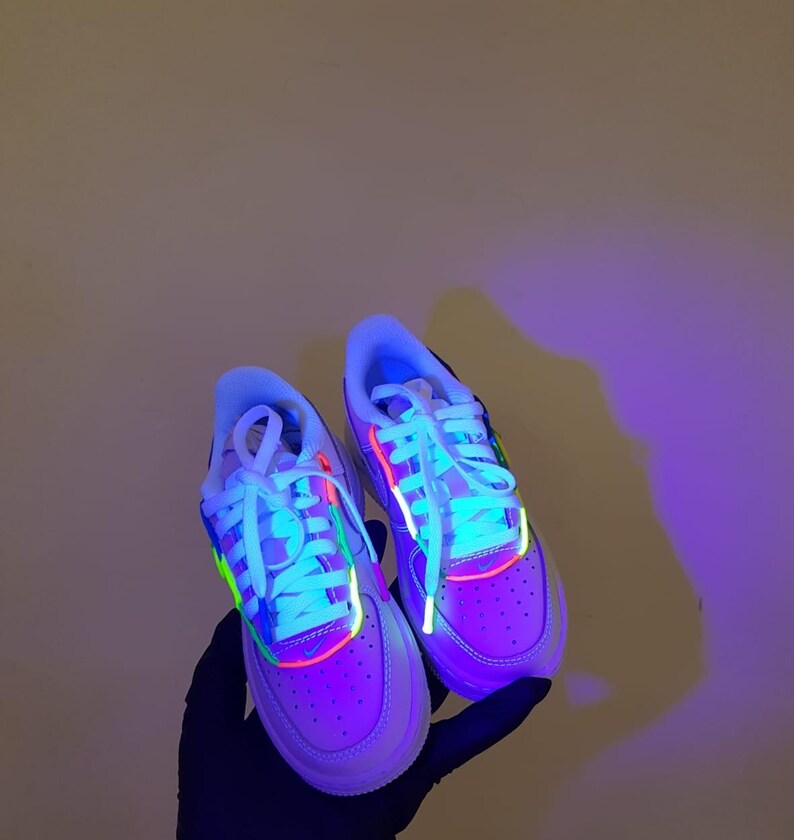 Puede incluir: Un par de zapatillas blancas con detalles en verde ne&oacute;n, rosa y azul. Las zapatillas est&aacute;n iluminadas con luz negra, lo que hace que los colores ne&oacute;n brillen intensamente.