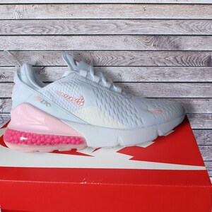 May include: Light blue Nike Air Max 270 athletic shoe with a pink bubble heel and pink accents. The shoe features a white sole and a pink Nike swoosh logo. The shoe is on a red box with a white Nike logo.