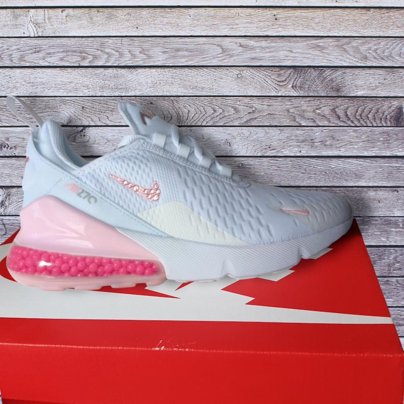 May include: Light blue Nike Air Max 270 athletic shoe with a pink bubble heel and pink accents. The shoe features a white sole and a pink Nike swoosh logo. The shoe is on a red box with a white Nike logo.