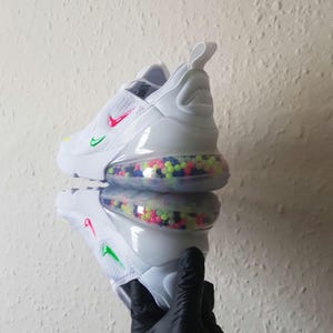 May include: White athletic shoe with a colorful bubble-filled sole. The shoe features a red and green swoosh logo and a pull tab. The shoe is reflected in a mirrored surface.