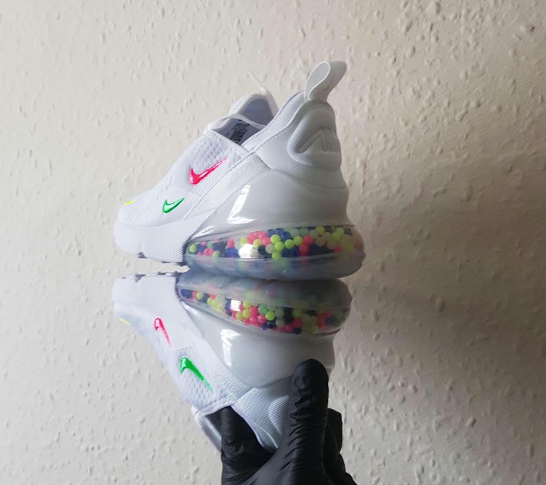 May include: White athletic shoe with a colorful bubble-filled sole. The shoe features a red and green swoosh logo and a pull tab. The shoe is reflected in a mirrored surface.