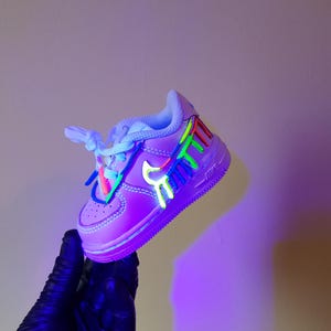 Puede incluir: Un par de zapatillas blancas con una base morada y salpicaduras de pintura de colores. Las zapatillas est&aacute;n iluminadas con luz negra, lo que hace que la pintura brille.