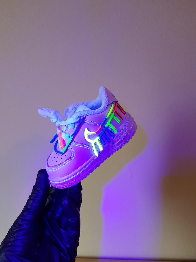 Puede incluir: Un par de zapatillas blancas con una base morada y salpicaduras de pintura de colores. Las zapatillas est&aacute;n iluminadas con luz negra, lo que hace que la pintura brille.