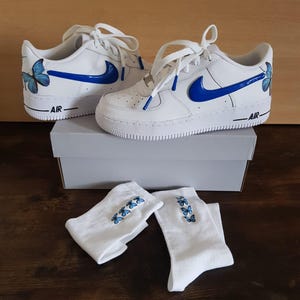 Może przedstawiać: Białe sk&oacute;rzane buty sportowe z niebieskimi wzorami motyli i niebieskim logo Swoosh. Buty są w komplecie z białymi skarpetkami, kt&oacute;re r&oacute;wnież mają niebieskie wzory motyli.