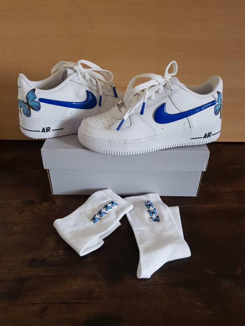 Może przedstawiać: Białe sk&oacute;rzane buty sportowe z niebieskimi wzorami motyli i niebieskim logo Swoosh. Buty są w komplecie z białymi skarpetkami, kt&oacute;re r&oacute;wnież mają niebieskie wzory motyli.