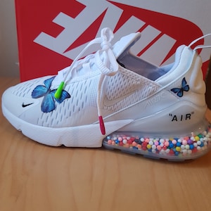 Puede incluir: Zapatillas Nike Air Max 270 blancas con un dise&ntilde;o de mariposa en el lateral y una suela transparente con chispas de colores. El texto "AIR" est&aacute; impreso en el lateral de la zapatilla.