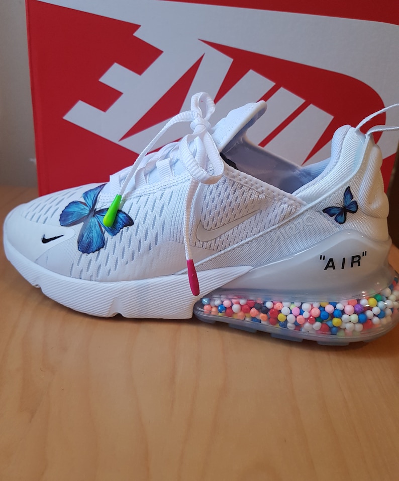 Puede incluir: Zapatillas Nike Air Max 270 blancas con un dise&ntilde;o de mariposa en el lateral y una suela transparente con chispas de colores. El texto "AIR" est&aacute; impreso en el lateral de la zapatilla.
