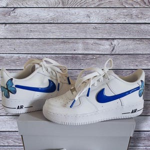 Może przedstawiać: Białe sk&oacute;rzane buty sportowe z niebieskimi wzorami motyli i swoosh. Buty mają napis "AIR" na boku.