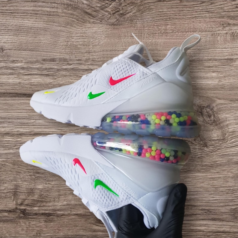 May include: White Nike Air Max 270 sneakers with red and green swoosh logos. The soles have a clear section filled with colorful beads. The shoes are on a wooden surface.