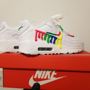 Op de afbeelding: Witte Nike Air Max 90 sneakers met een kleurrijk verfspat-ontwerp aan de zijkant. De schoenen staan op een rode Nike schoenendoos.