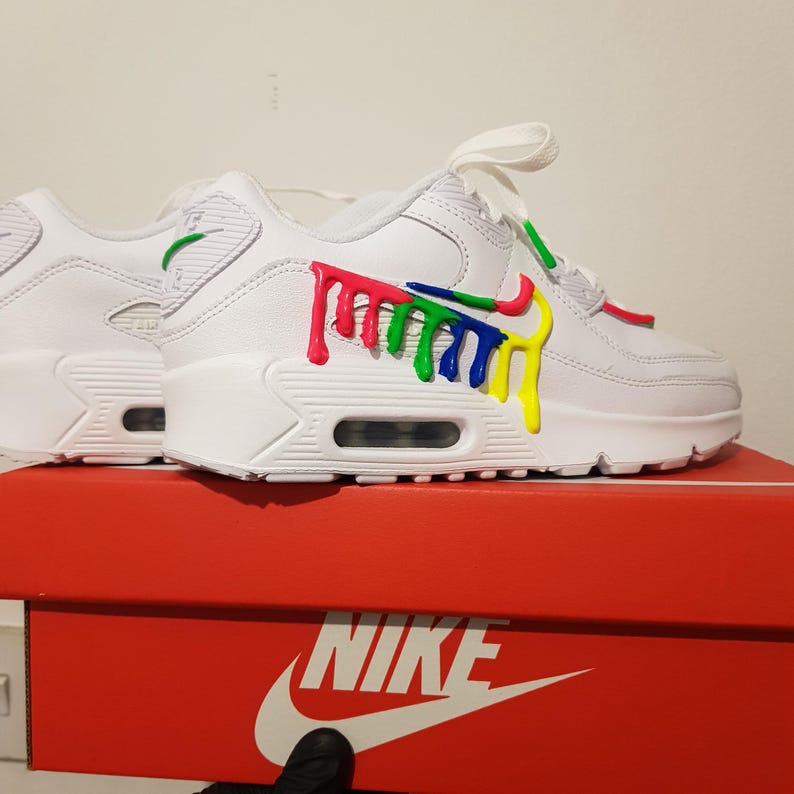 Op de afbeelding: Witte Nike Air Max 90 sneakers met een kleurrijk verfspat-ontwerp aan de zijkant. De schoenen staan op een rode Nike schoenendoos.