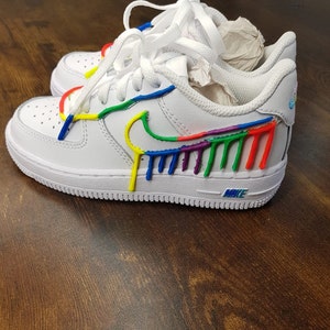 Puede incluir: Zapatillas bajas de cuero blanco con un dise&ntilde;o de pintura arco&iacute;ris en el swoosh y el lateral de la zapatilla. El texto "NIKE" es visible en la suela.