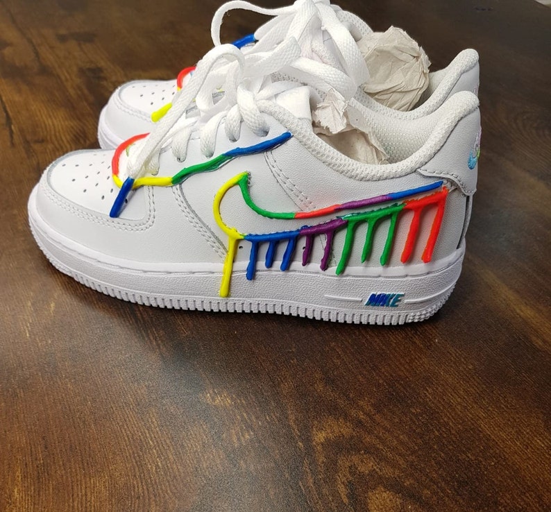 Puede incluir: Zapatillas bajas de cuero blanco con un dise&ntilde;o de pintura arco&iacute;ris en el swoosh y el lateral de la zapatilla. El texto "NIKE" es visible en la suela.