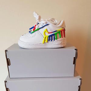 Puede incluir: Zapatillas blancas de cuero con un dise&ntilde;o de salpicaduras de pintura de colores en el lateral. Las zapatillas est&aacute;n encima de dos cajas blancas.