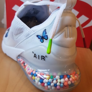 Puede incluir: Zapatillas Nike Air Max 270 blancas con un gr&aacute;fico de mariposa azul en la parte posterior. El zapato tiene una suela de burbuja transparente llena de cuentas de colores y la palabra "AIR" impresa en la parte posterior.
