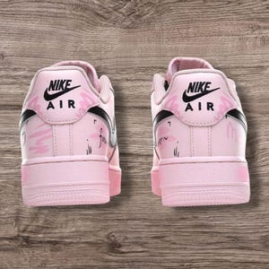 May include: Pair of pink Nike Air Force 1 sneakers. The shoes have a light pink base with black accents and the Nike logo. The back of the shoes feature a floral design and the word "forever" is written on the side.