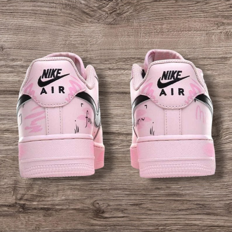 May include: Pair of pink Nike Air Force 1 sneakers. The shoes have a light pink base with black accents and the Nike logo. The back of the shoes feature a floral design and the word "forever" is written on the side.