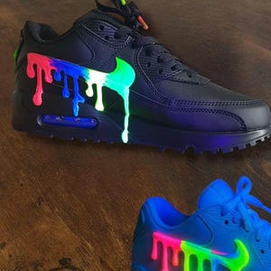 May include: Black sneakers with a colorful paint drip design. The Nike swoosh and the dripping paint are in bright green, white, blue, and red. The laces have matching colorful accents. The second sneaker is blue with a similar design.