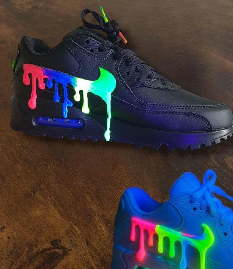 May include: Black sneakers with a colorful paint drip design. The Nike swoosh and the dripping paint are in bright green, white, blue, and red. The laces have matching colorful accents. The second sneaker is blue with a similar design.