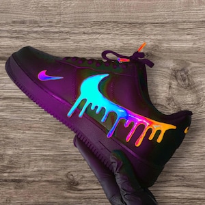 May include: Black Air Force 1 sneaker with a vibrant, iridescent drip design. The Nike swoosh is a bright blue, and the laces have orange and purple accents. The shoe is held by a gloved hand.