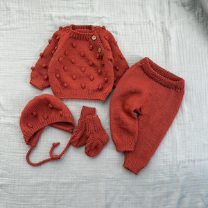 May include: A knitted baby clothing set in a warm orange-red colour. The set includes a sweater with bobble detailing, trousers, a bonnet, and socks. The sweater has wooden buttons. The items are arranged on a light grey fabric.