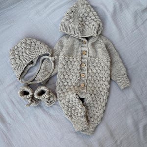 May include: A light beige knitted baby outfit set, including a hooded jumpsuit with wooden buttons, a bonnet, and booties. The set features a textured diamond pattern and is made of soft yarn.