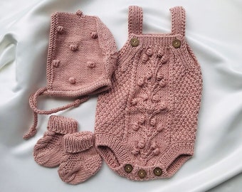Könnte beinhalten: Ein rosa gestricktes Baby-Outfit-Set mit Mütze, Stiefeln und einem Einteiler. Das Outfit ist aus strukturiertem Strick mit kleinen, erhabenen Noppen.