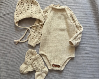 May include: A cream-colored knitted set consisting of a long-sleeved bodysuit with three brown buttons at the crotch, a matching bonnet with tie strings, and a pair of socks. The sleeves of the bodysuit, the bonnet, and the socks feature a delicate openwork pattern.